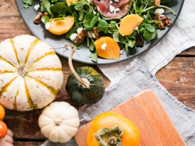 Persimmons and Watercress Salad with Candied Walnuts and Goat's Cheese on a black plate with Fall gourds and persimmons nearby.