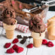 Dark Chocolate Raspberry Ice Cream with 2 cones with scoops of ice cream and chocolate and raspberries scattered around.