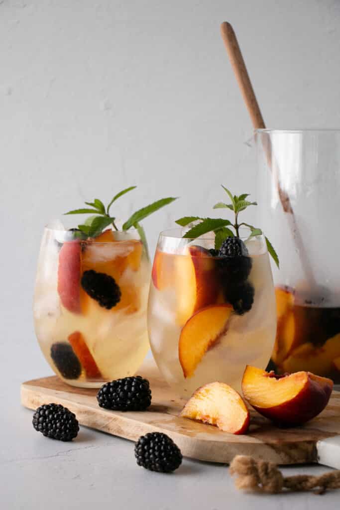 Two stemless wine glasses filled with ice, a white wine based sangria, peaches, blackberries and a bright green fresh mint stem. Blackberries and sliced peaches surround glasses and cutting board.