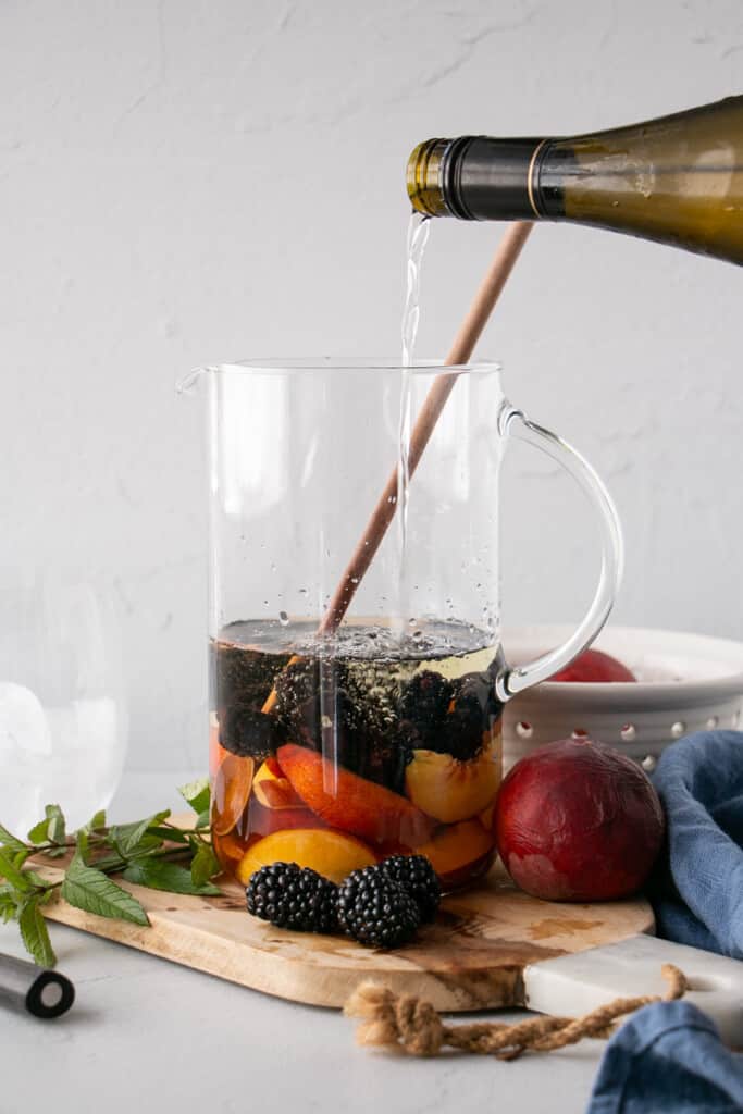 White wine bring poured into a clear modern jug containing blackberries and peaches for a Summer Sangria recipe.