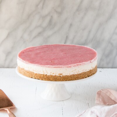 Side view a of No Bake Rhubarb Cheesecake on a white cake stand with a pink tea towel and a serving knife.