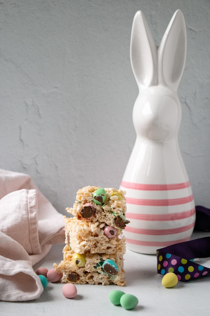 Easter Rice Krispie Treats piled 3 high with a white and pink striped ceramic bunny in the background. 