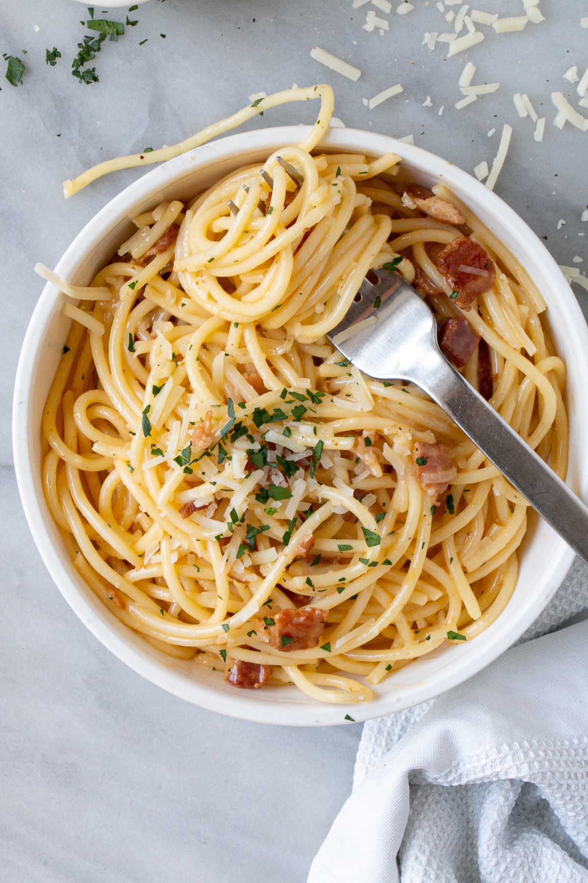 The best Spaghetti Carbonara in a white bowl with parmesan shreds and chopped parsley in small bowls and sprinkled on top.