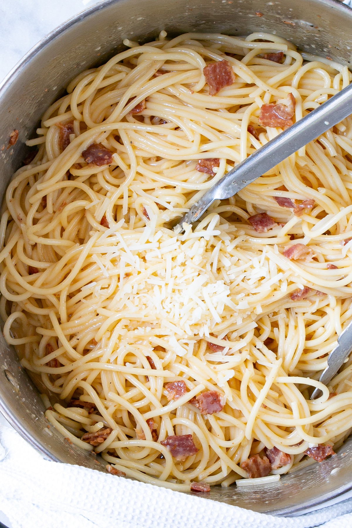 Spaghetti Carbonara in a large metal pot with metal tongs and parmesan cheese on top