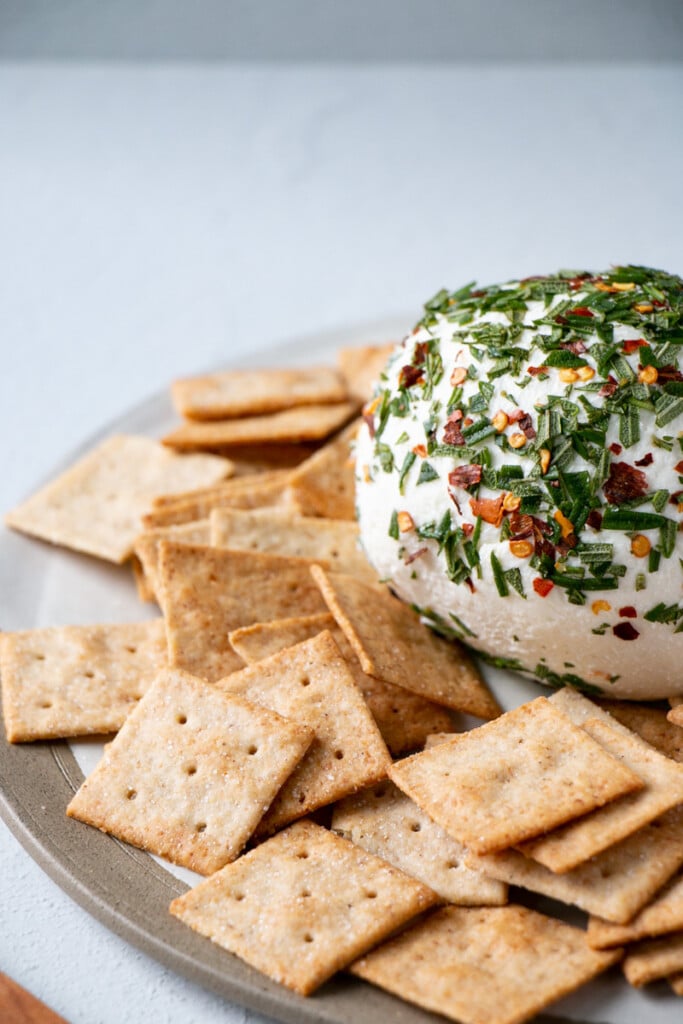 Honey Goat's Cheese Ball recipe coated in chopped fresh rosemary and crushed red pepper flakes with square almond crackers surrounding it. 