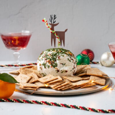 Honey Goat's Cheese Ball with chopped rosemary and crushed pepper flakes on the exterior with cracker on a plate and festive decor around.