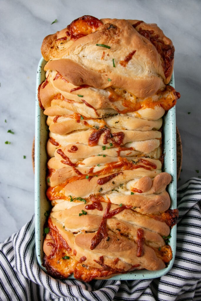 Pull Apart Cheese Bread in a teal loaf pan with melted cheddar cheese on top and a scattering of chopped parsley.