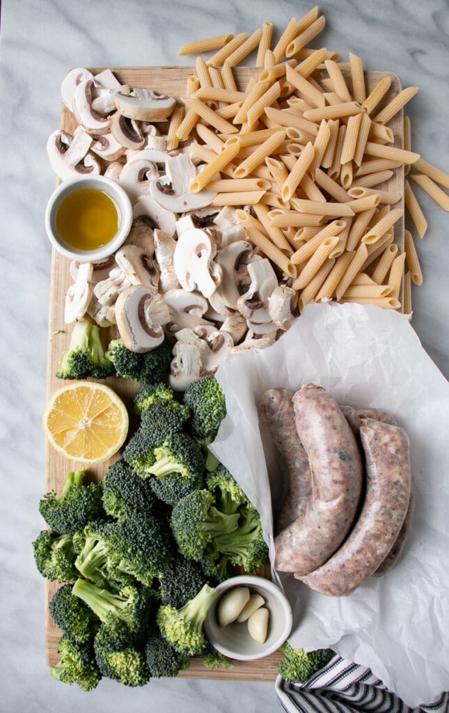 Raw ingredients on a cutting board including: Italian Sausage, Broccoli, Mushrooms, Penne Pasta, 1/2 cut lemon, olive oil, and garlic cloves.