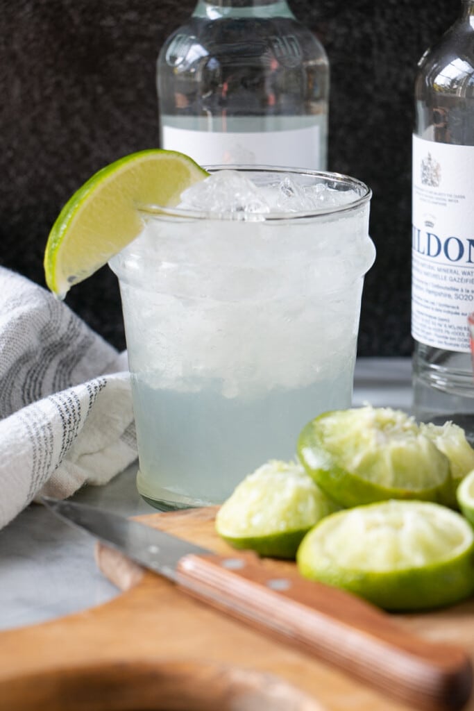 A glass of ice, tequila, lime juice and crushed ice with a slice of lime on the edge, surrounded by squeezed limes.