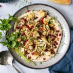 Crispy Cauliflower on a bed of smooth hummus and topped with pomegranate seeds and chopped parsley on a grey ceramic plate and small bunch of parsley stems on the side of the dish.